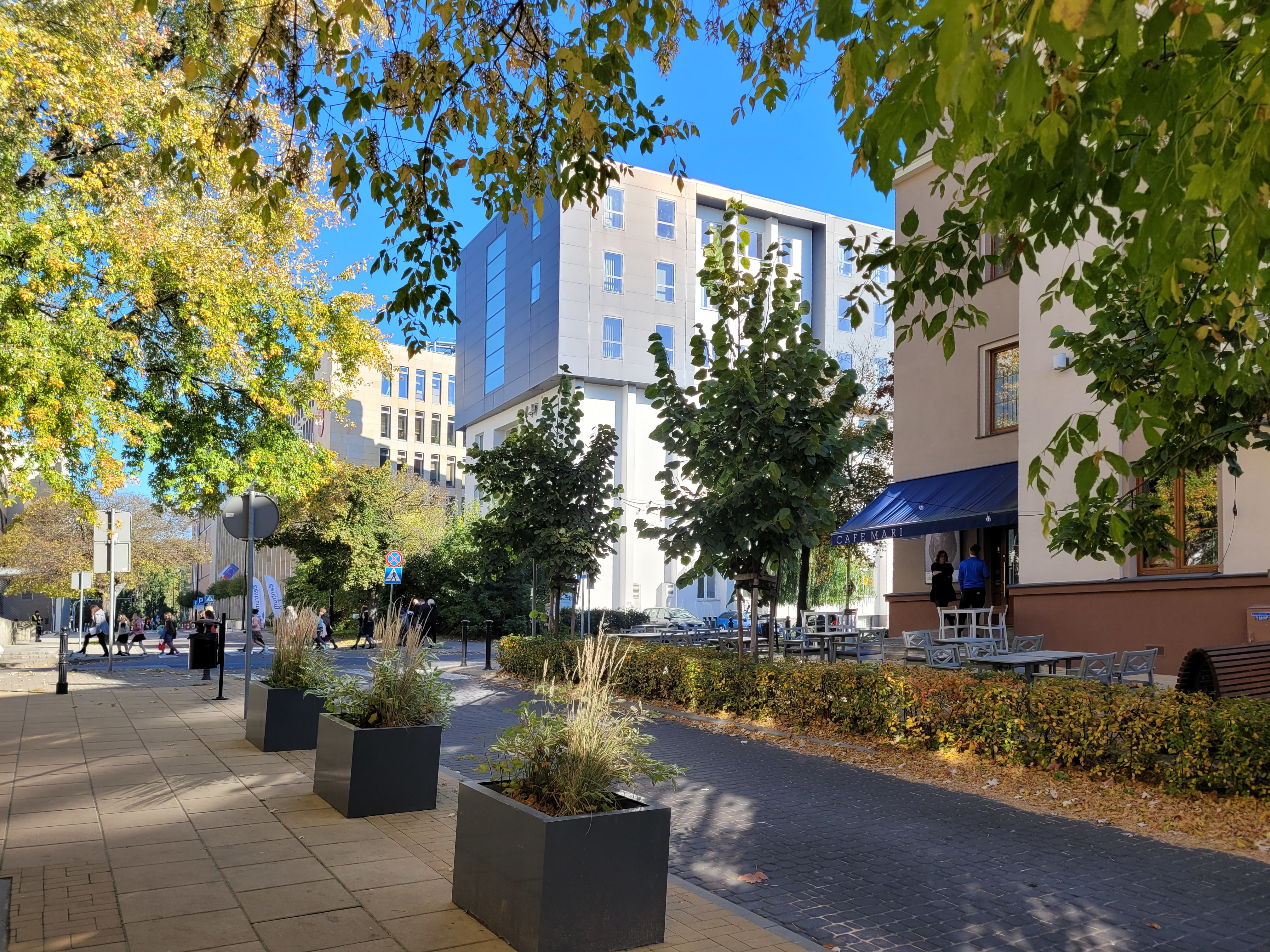 Miejska ulica z chodnikiem, roślinami w donicach i drzewami; po prawej stronie znajduje się budynek z ogr&oacute;dkiem kawiarnianym, a w tle widać nowoczesne biurowce.